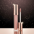 A tube of glittery rose gold lip gloss stands next to its applicator wand on a reflective surface against a white background with subtle gold speckles.